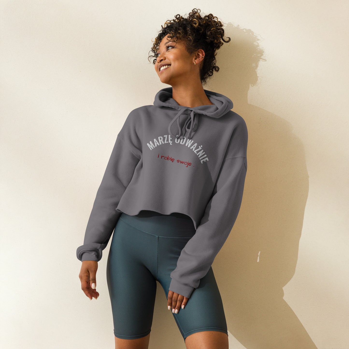 Ein Model trägt den grauen Cropped Hoodie von POLCOUTURE. Die Frontansicht zeigt den modernen, bauchfreien Schnitt und die Stickerei „słucham serca" in weißt, sowie "ono szepcze“ in rot. Das Model kombiniert den Hoodie lässig mit einer kurzen Sporthose.