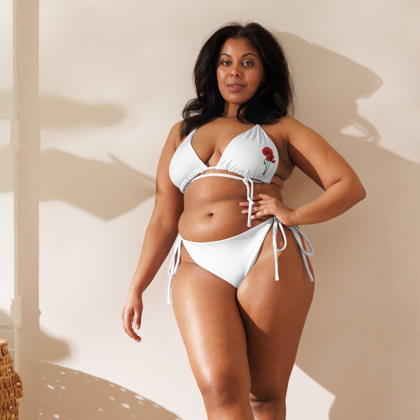 Ein kurviges Model lehnt an einer Wand und trägt den weißen Bikini „MAKI“ von POLCOUTURE. Fokus auf die vordere Passform und das florale Mohnblumen-Design.