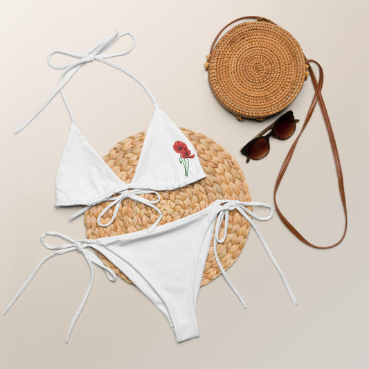 Weißer Bikini „MAKI“ von POLCOUTURE, flach arrangiert mit passenden Accessoires im Hintergrund. Das Design zeigt die charakteristische rote Mohnblume.