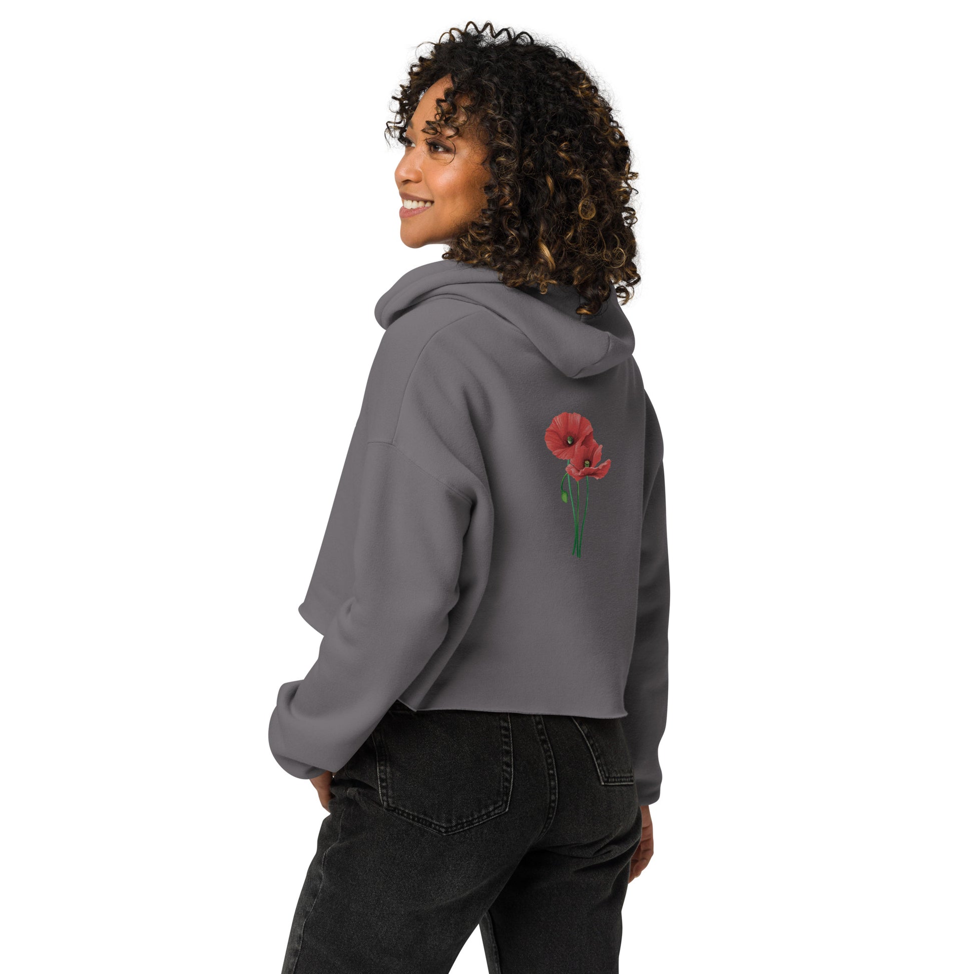 Seiten- und Rückenansicht eines Models mit braunen Locken, die den grauen Cropped Hoodie von POLCOUTURE trägt. Das Model blickt über die Schulter zur Seite, wodurch der minimalistische Mohnblumen-Print auf dem Rücken sowie der lockere, bauchfreie Schnitt des Hoodies perfekt zur Geltung kommen.