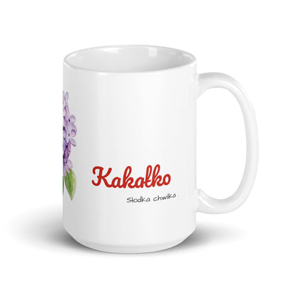 Seitenansicht der weißen POLCOUTURE Tasse mit Henkel rechts. Im Fokus steht das polnische Wort „kakałko“, mit dem Slogan „słodka chwilka“ klein direkt darunter. Die fliederfarbenen Blüten sind bei dieser Ansicht kaum zu sehen.